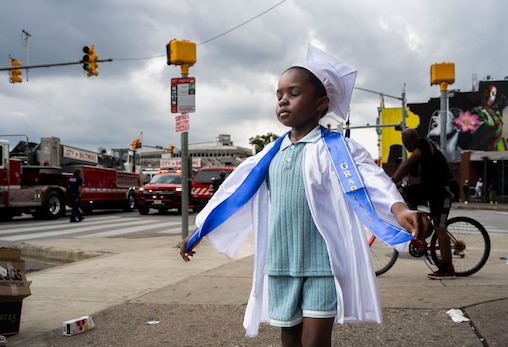 Taylin Lawrence, 5, soaks in the excitement of graduating from preschool. He says he wants to grow up to be a cop because they help people. In the background, first responders handle an alleged mass overdose situation involving fifteen people at the intersection of Pennsylvania and North Avenues.