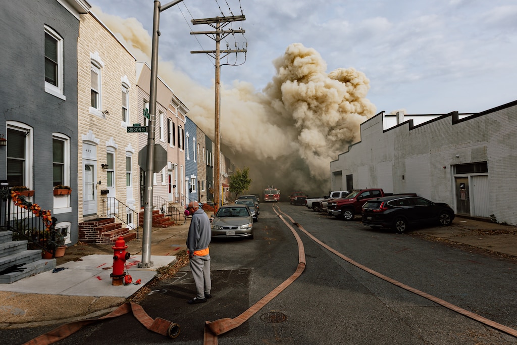 A pedestrian stands by while a three-alarm fire blazes at a commercial building in Remington, in Baltimore, MD on Friday, Nov. 7, 2025.