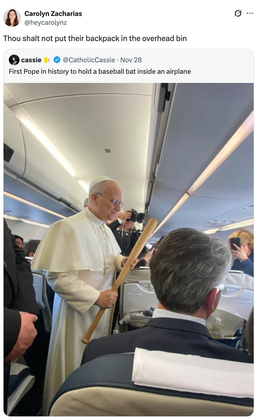 A person dressed in papal attire humorously holds a large baseball bat in an airplane aisle, amusing passengers onboard