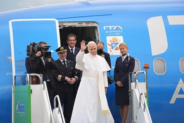 A religious leader in traditional attire waves while exiting a plane, accompanied by airline staff and photographers capturing the moment