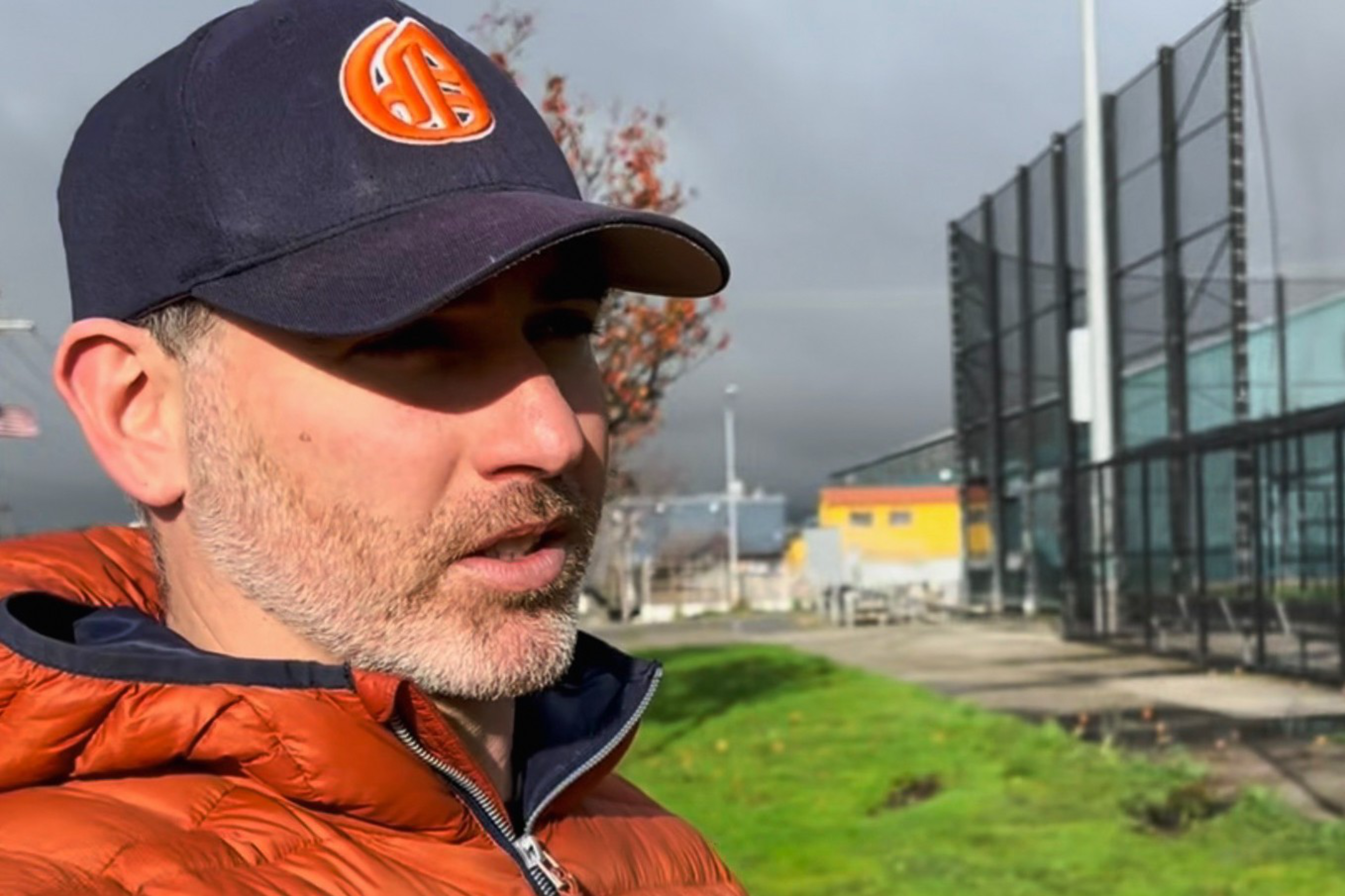 A man with stubble wears a navy cap and orange puffer jacket, standing near a fenced outdoor sports area under a cloudy sky.