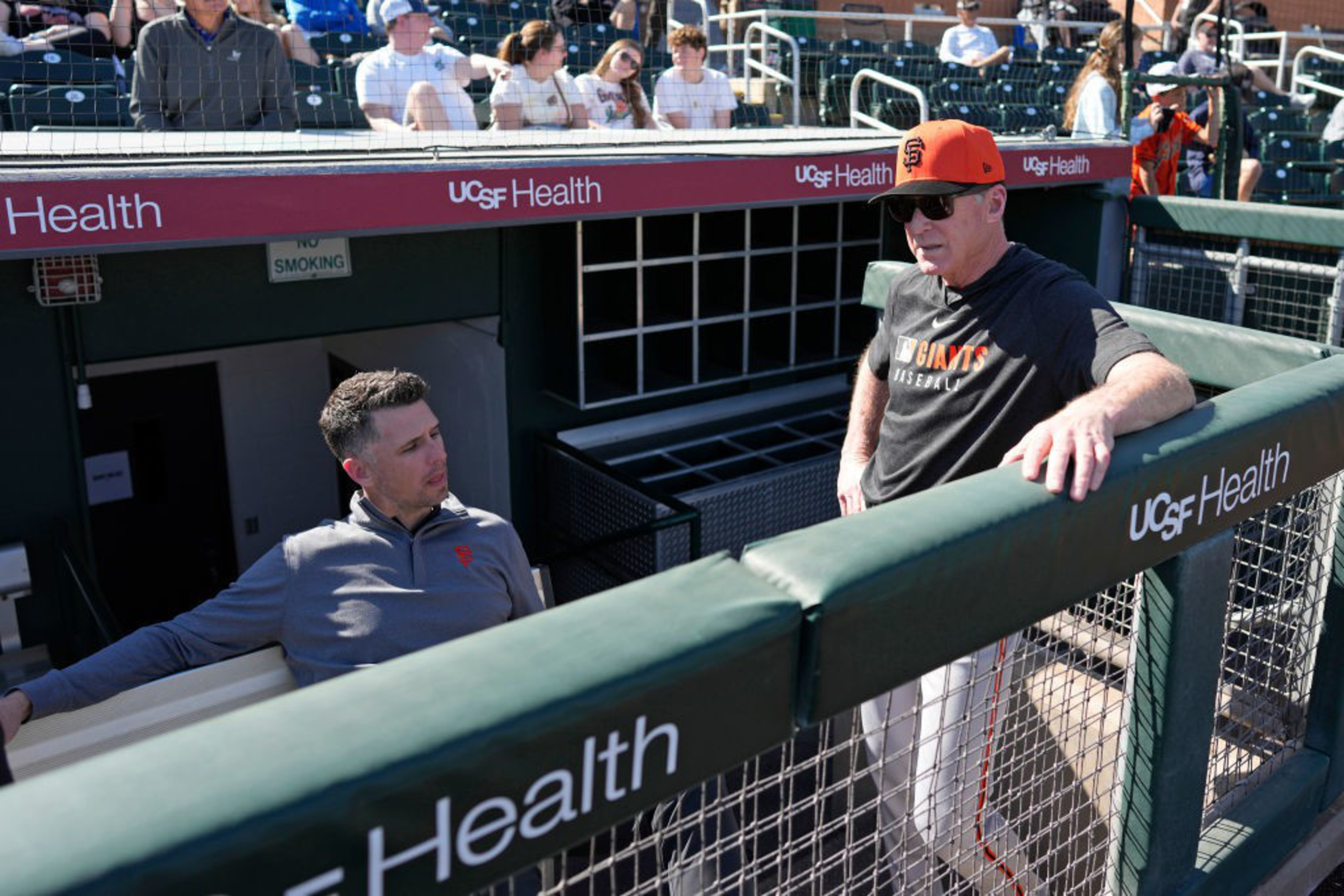 Two men in a dugout are talking casually. One is seated, wearing a gray jacket, and the other, standing, wears a black “Giants Baseball” shirt and cap.