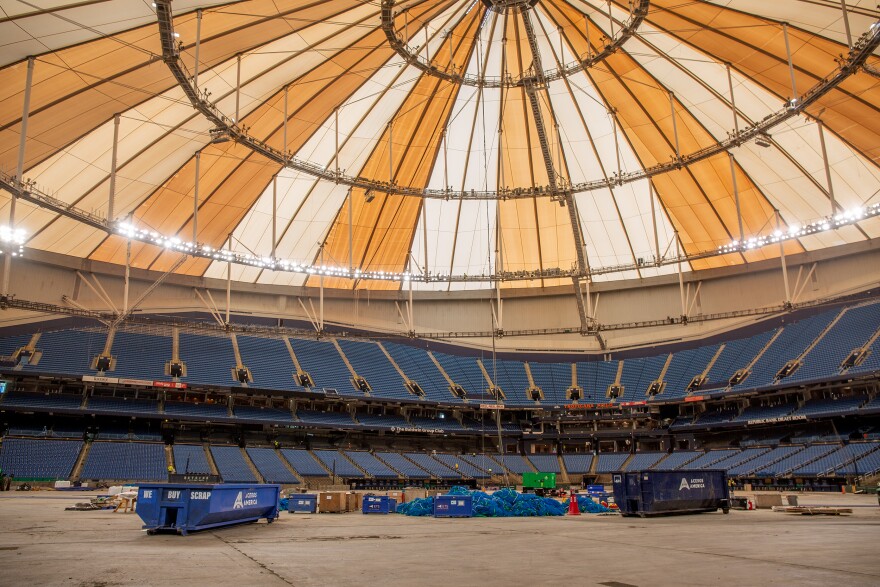 Inside Tropicana Field. Brown and white triangular roof panels are shown over the blue stadium seating and bare flooring where the baseball field resides. 