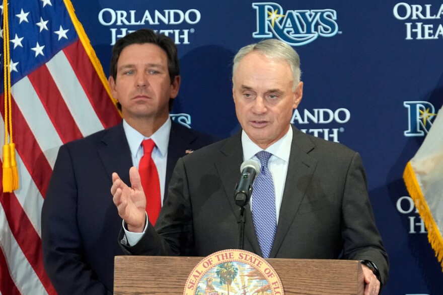 Man in a blue suit to the left, behind a man in a gray suit talking at a podium with a U.S. flag to the left and in front of a blue backdrop