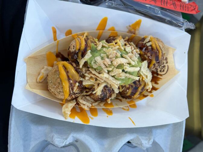 A wooden tray resembling a small boat holds fried balls drenched in orange and brown sauce. In the middle sits a generous dollop of guacamole topped with crispy garnishes.