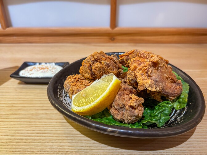 Various pieces of fried chicken and a slice of lemon sit inside a small brown dish. Behind it in the background is a white dipping sauce. 