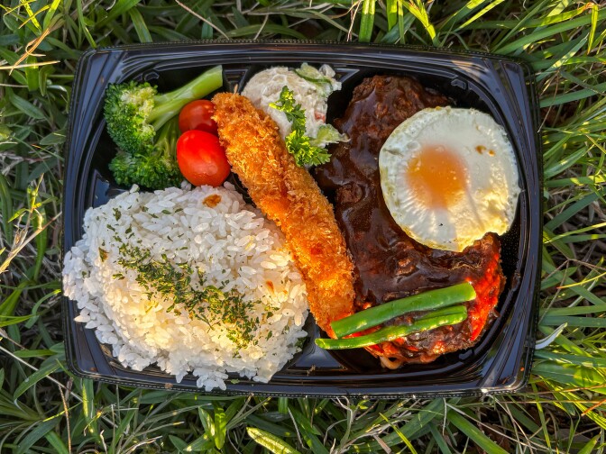 A black plastic tray sits against a green grass backdrop. It is filled with cooked white rice, a light golden brown color fried chicken tender, and a large brown steak with a small circular fried egg. 