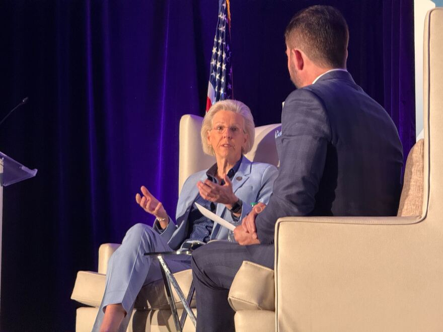 Woman in a light blue suit with blonde hair and glasses gestures while sitting in a chair. A man sitting in another chair is facing her. An American flag is behind her. 