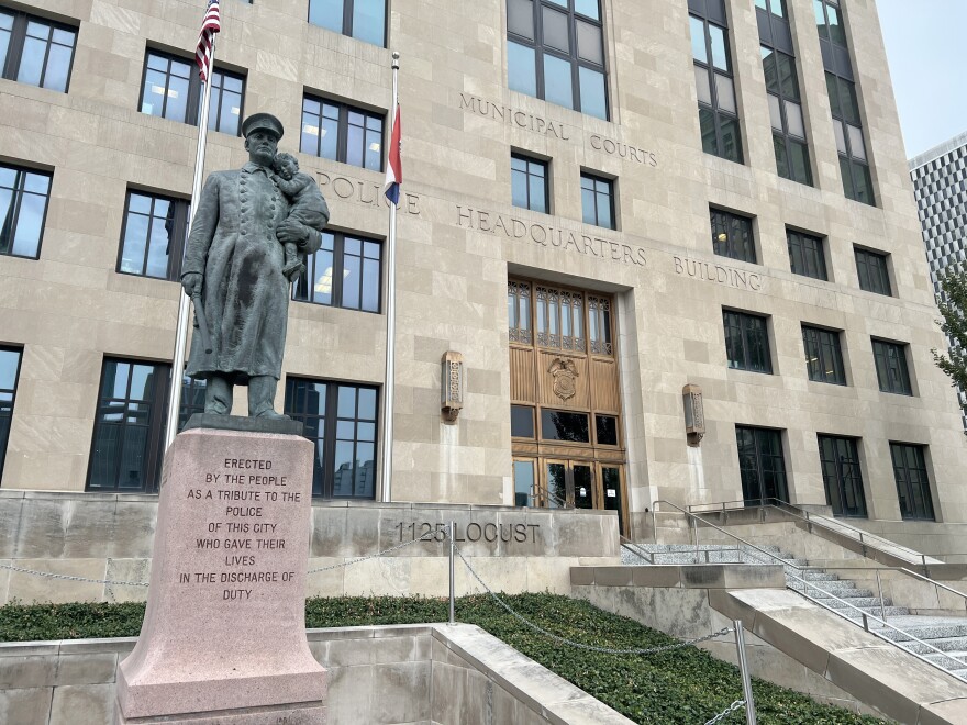 A bronze statue of a police officer is shown on a granite pedestal  in front of a large stone building with the carved letters in the building that read "Municipal Court, Police Headquarters Building."