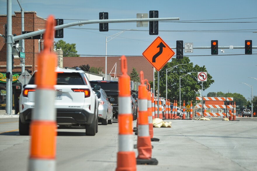 Traffic pylons lines the middle of a freshly poured roadway where traffic can be seen driving on one side of the pylons. There are more pylons and detour barriers in the background at an intersection where the traffic lights have turned red.