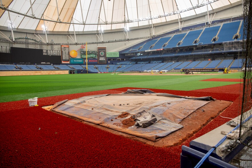 A look at ground level across an indoor baseball stadium. A rust-colored artifical grass surrounds the brown dirt on home plate. You can see blue stadium seating in the background.