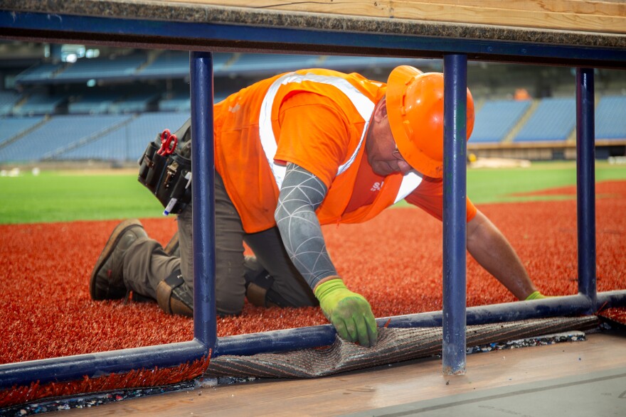 A close up of a man in a yellow safety vest and hardhat on his hands and knees. He's laying down rust colored artificial grass on the outer edge of the field.