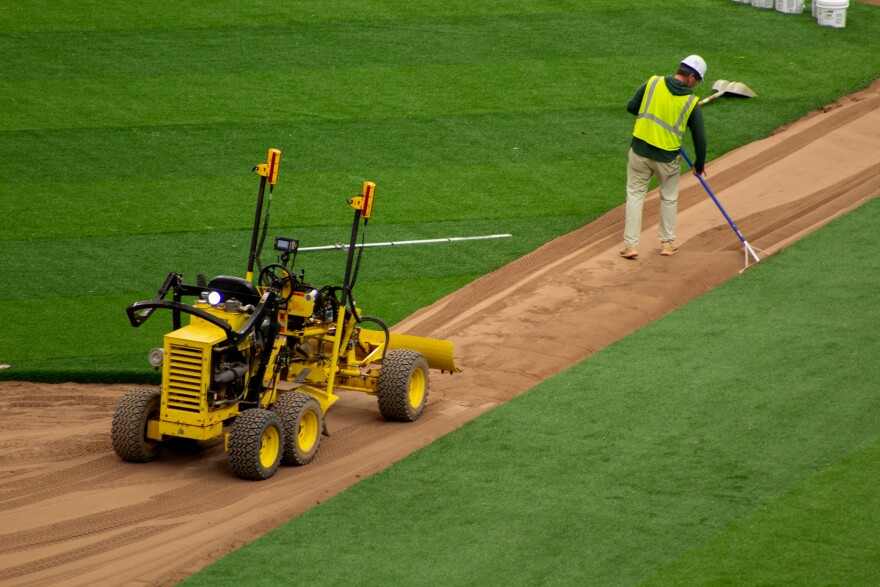 A man wearing a yellow safety vest and white hardhat is raking dirt on the field. A few feet behind him is a small yellow and black construction vehicle.
