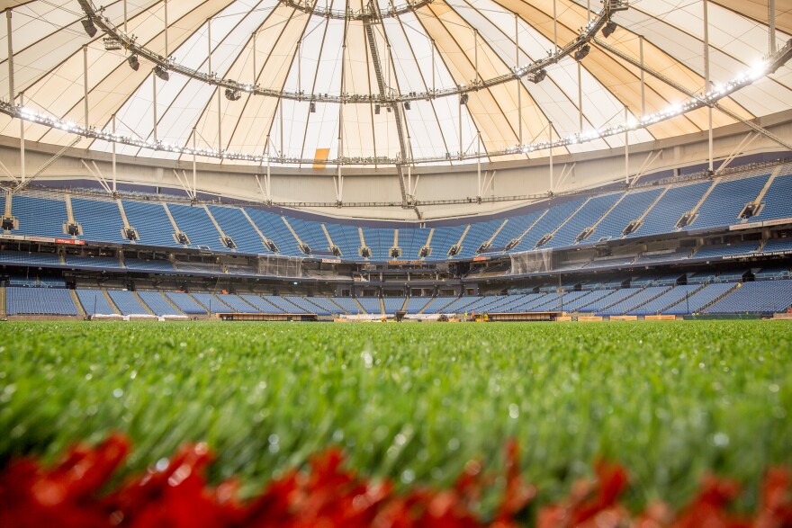 Shot from the ground. In the foreground and blurred out is rust and green colored artificial grass. In the backround, in focus, is the rest of the indoor baseball field with blue stadium seating and a peek at the beige and white triangular fiberglass roof panels covering it all.