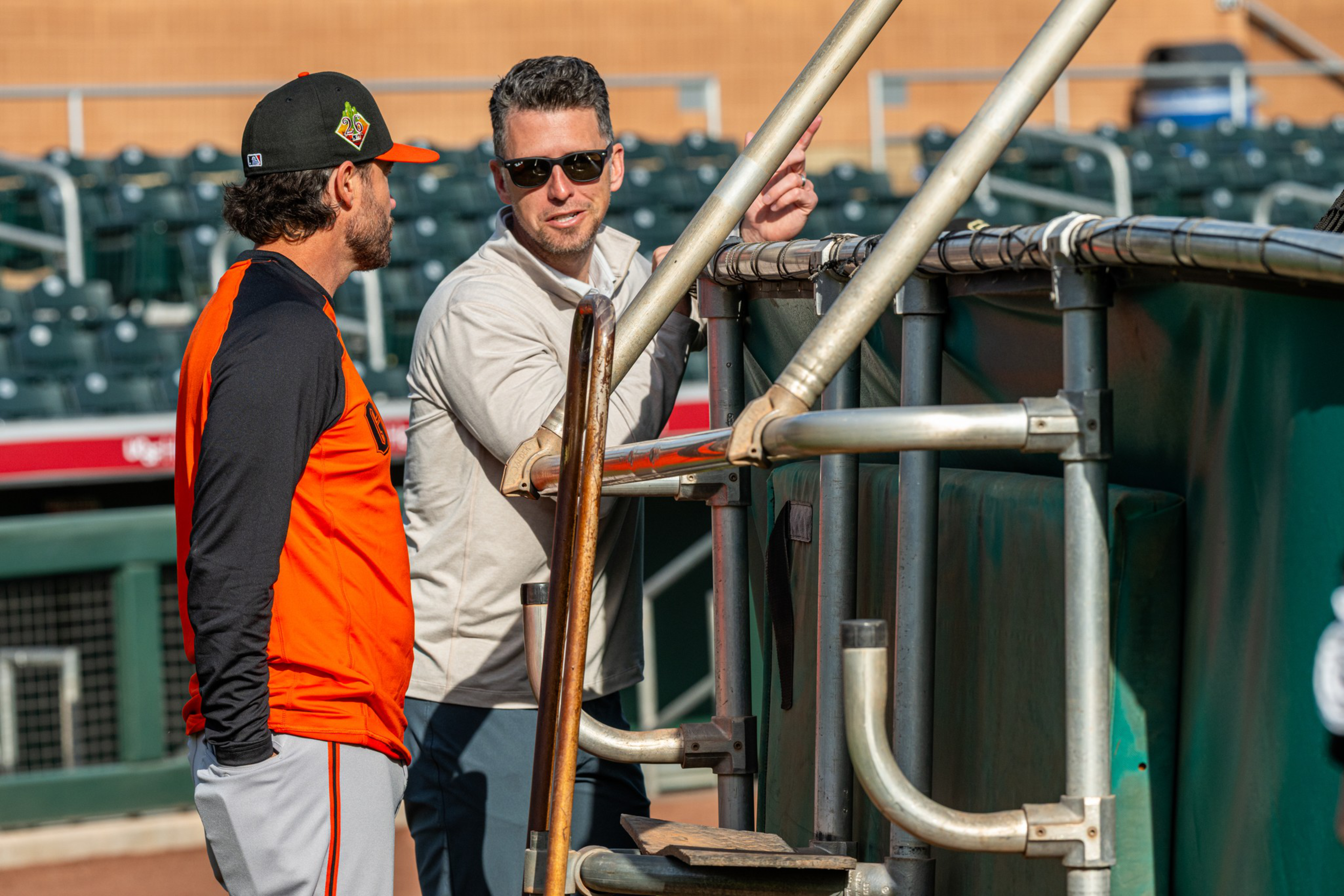 Two men, one in an orange and black sports outfit and the other in sunglasses and a light shirt, are talking near a baseball dugout.