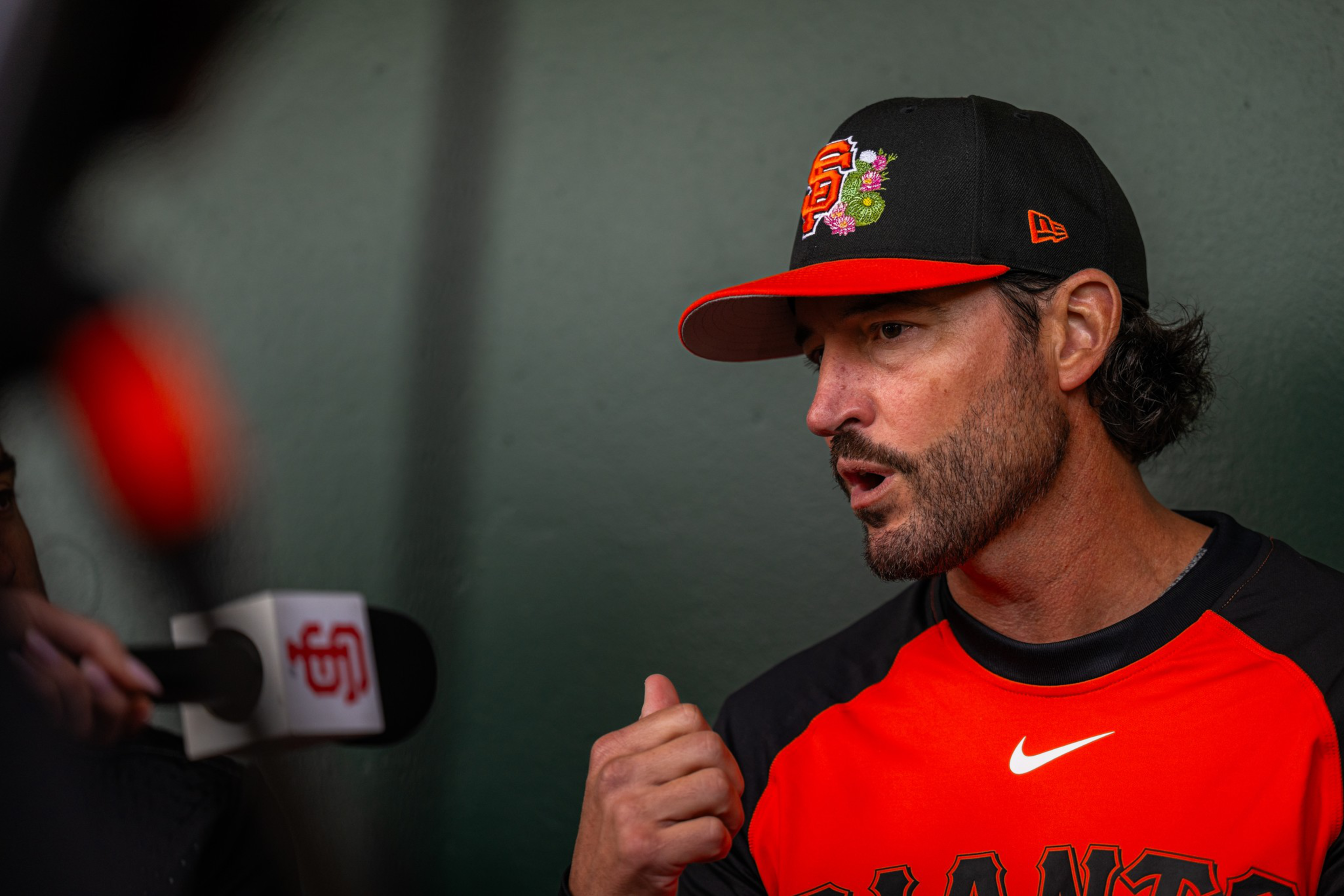 A man wearing a San Francisco Giants cap and jersey is speaking, gesturing with his hand, while a microphone labeled “sf” is held nearby.