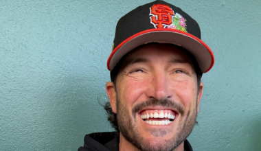 A man with a beard is smiling broadly, wearing a black and orange San Francisco Giants baseball cap and a black hoodie.