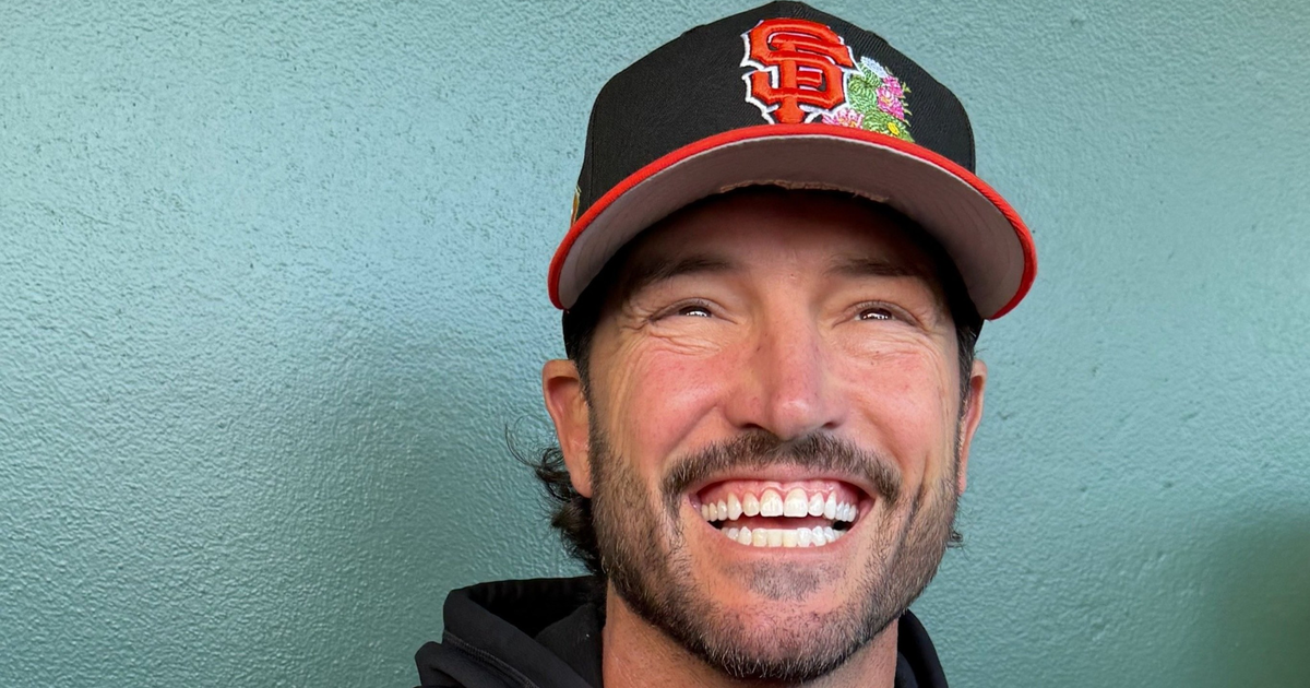 A man with a beard is smiling broadly, wearing a black and orange San Francisco Giants baseball cap and a black hoodie.