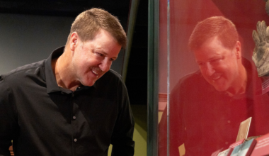 A man in a black shirt leans forward, smiling, as he looks at historical documents and a glove displayed in a glass case.