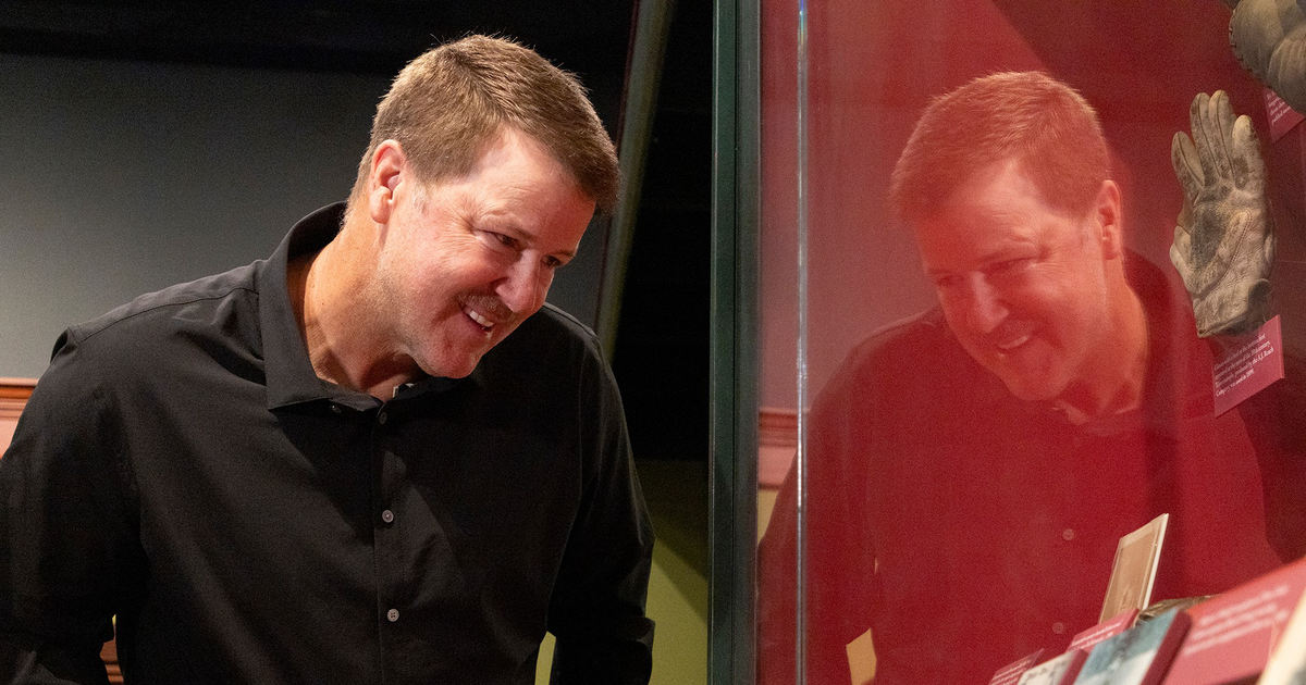 A man in a black shirt leans forward, smiling, as he looks at historical documents and a glove displayed in a glass case.