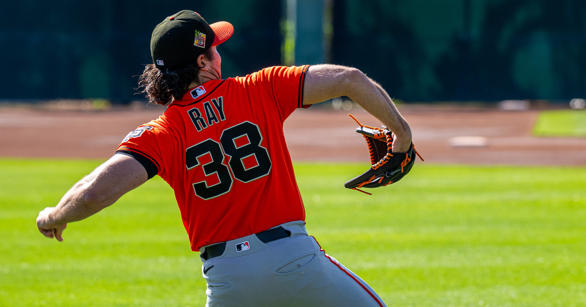 A baseball pitcher wearing an orange jersey with "RAY 38" on the back is in mid-throw on a grassy field.