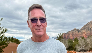 A man wearing a light gray sweater and dark sunglasses stands near a road with trees and red rock mountains in the cloudy background.