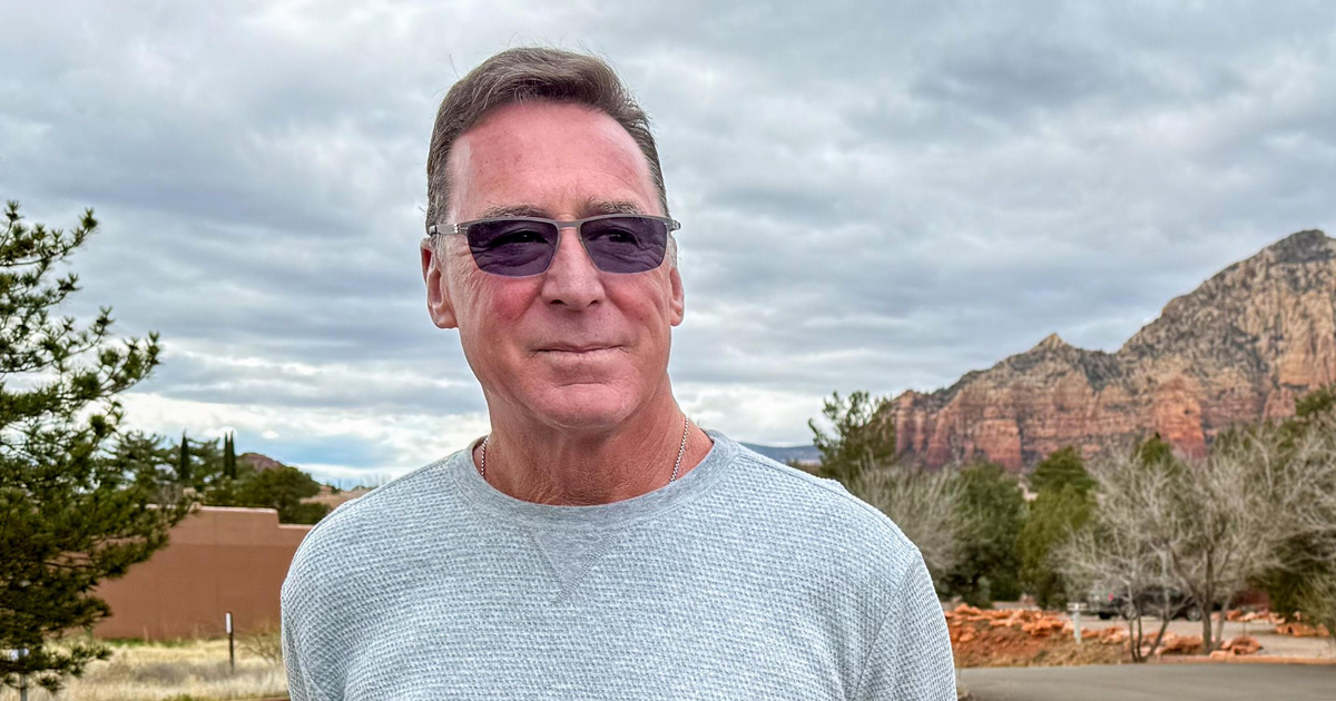 A man wearing a light gray sweater and dark sunglasses stands near a road with trees and red rock mountains in the cloudy background.
