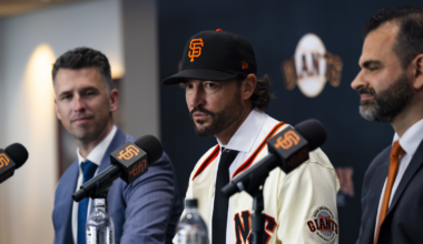 Three men sit at a table with microphones; the center man wears a San Francisco Giants baseball cap and jersey, flanked by two men in suits.