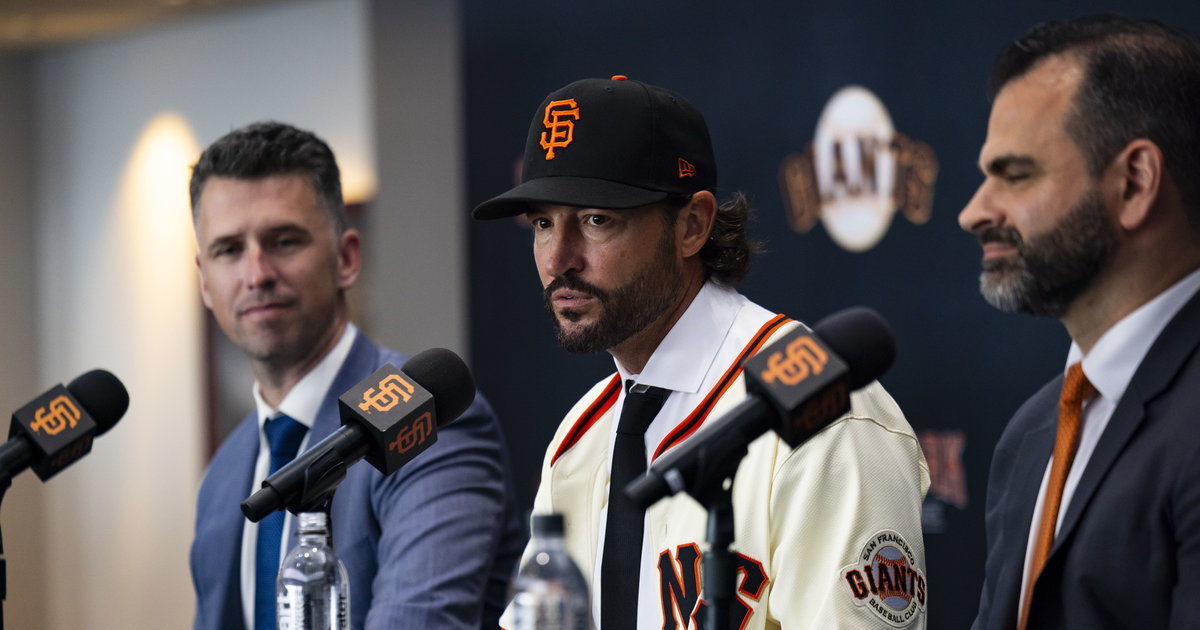 Three men sit at a table with microphones; the center man wears a San Francisco Giants baseball cap and jersey, flanked by two men in suits.