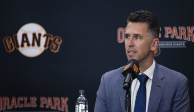A man in a blue suit and tie speaks at a press conference with a Giants logo and Oracle Park backdrop behind him.