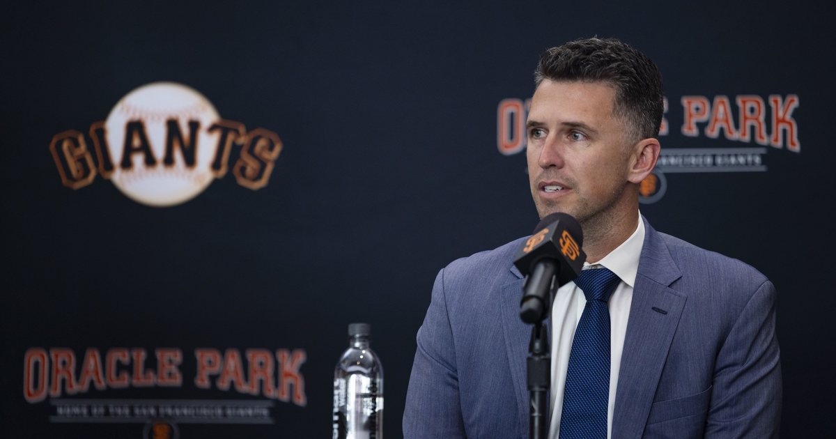 A man in a blue suit and tie speaks at a press conference with a Giants logo and Oracle Park backdrop behind him.