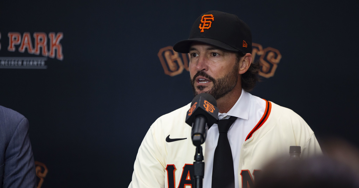 A man wearing a San Francisco Giants baseball jersey and cap speaks into a microphone at a press conference.