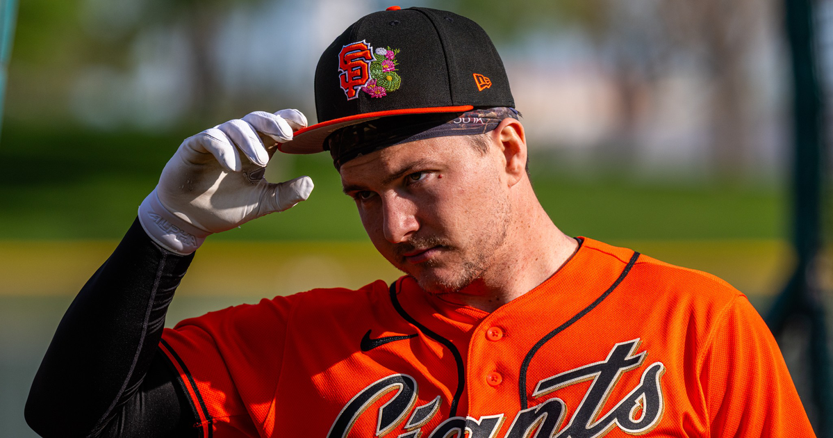 A baseball player in an orange Giants jersey adjusts a black and orange cap with the team's logo and floral design, wearing a white glove on one hand.