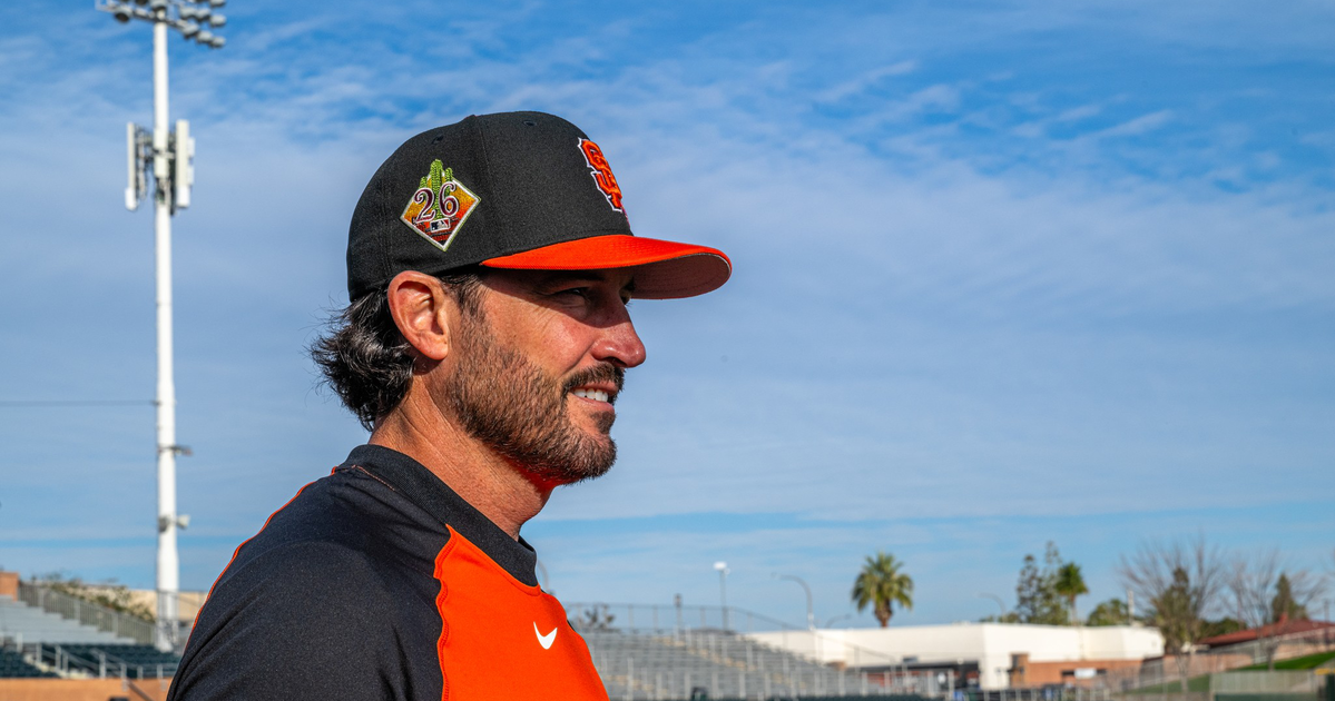 A man with a beard wearing a San Francisco Giants baseball cap and jersey stands on a baseball field under a partly cloudy sky.
