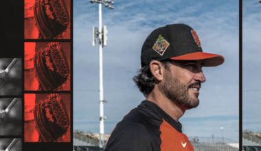 A baseball player wearing a black and orange Giants uniform faces right on a field with stadium lights and bleachers behind him.