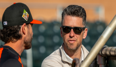 A man in sunglasses and a light jacket leans on a railing, talking to another man in a black and orange baseball uniform with a cap.