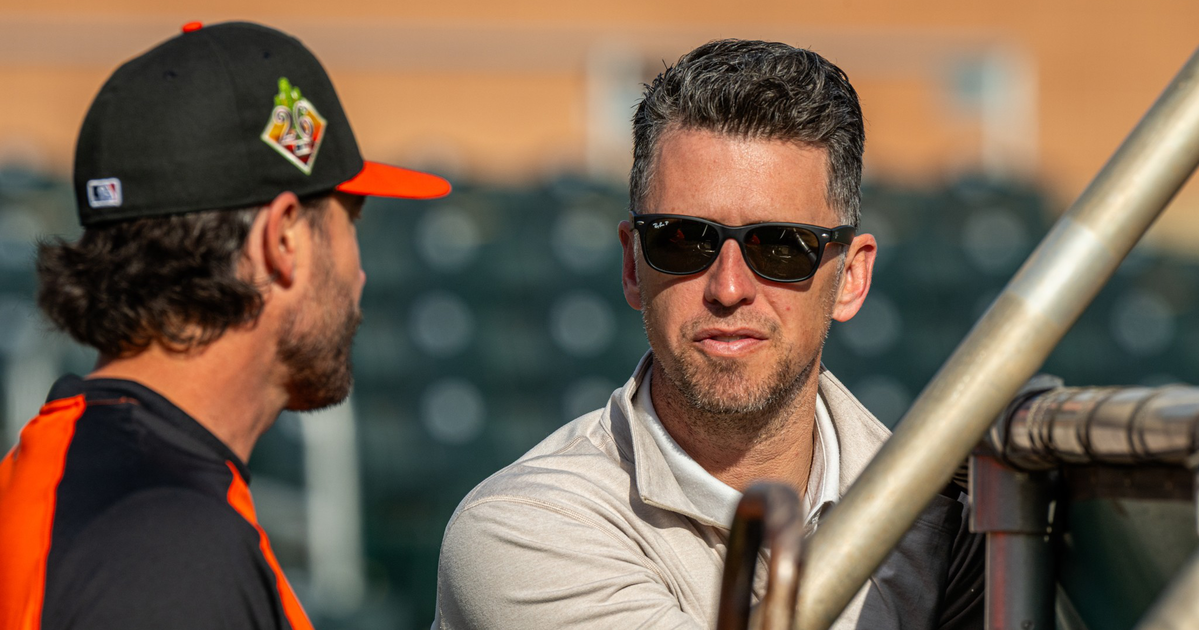A man in sunglasses and a light jacket leans on a railing, talking to another man in a black and orange baseball uniform with a cap.