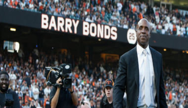 Barry Bonds in a black suit and white tie stands on a baseball field with a large crowd behind him and his name displayed on a scoreboard.