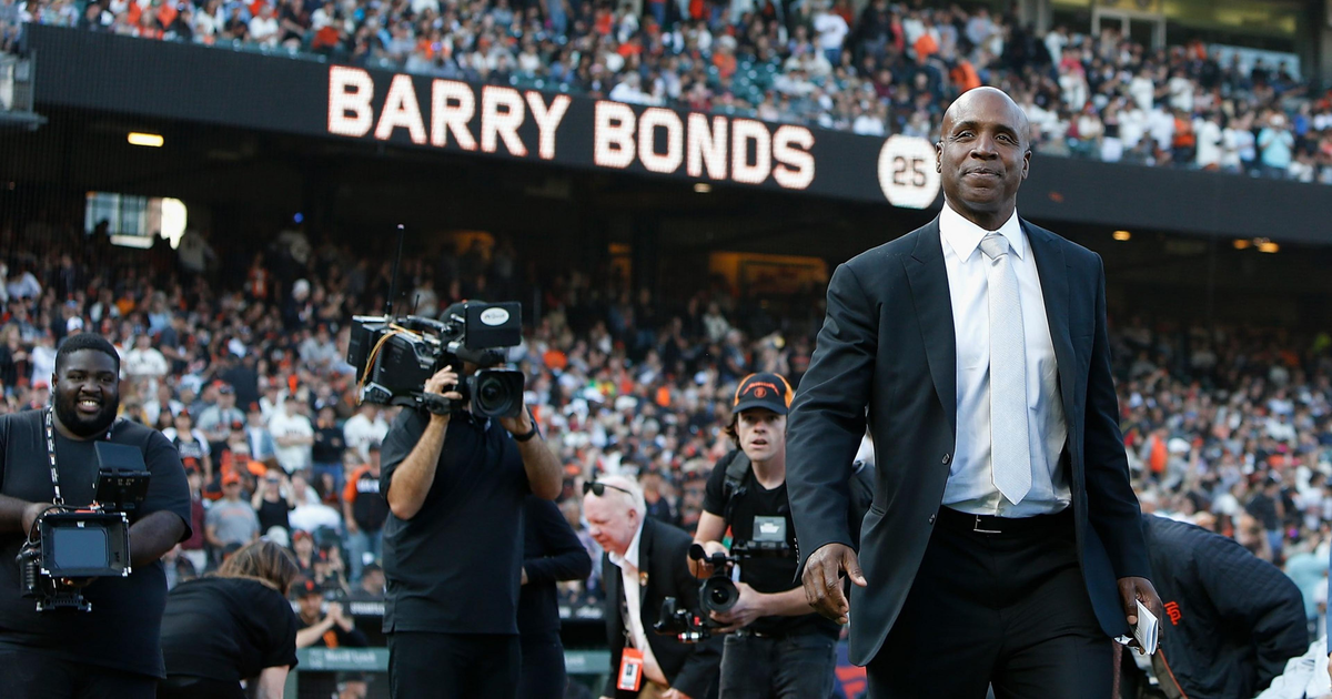 Barry Bonds in a black suit and white tie stands on a baseball field with a large crowd behind him and his name displayed on a scoreboard.