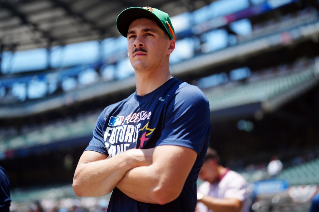 A male baseball player with a mustache, wearing a green baseball cap and a navy blue "All-Star Futures Game" t-shirt, stands with his arms crossed.