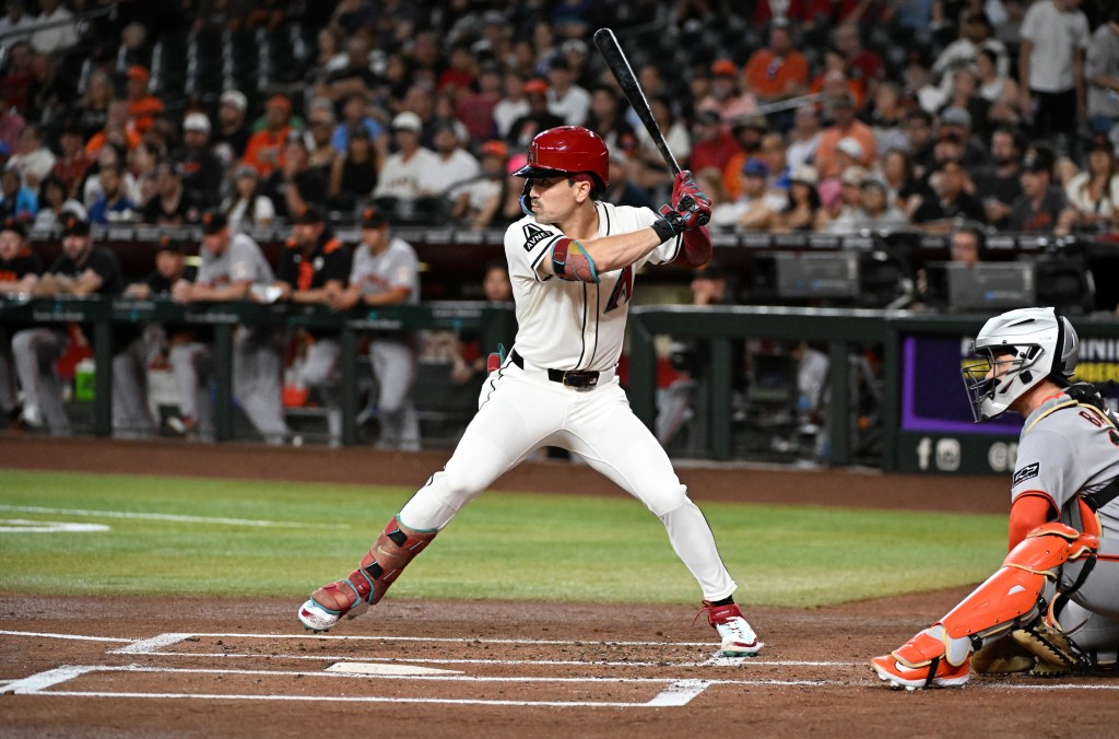 A baseball player in a red helmet and white uniform batting against the San Francisco Giants, with a catcher in orange gear behind him.