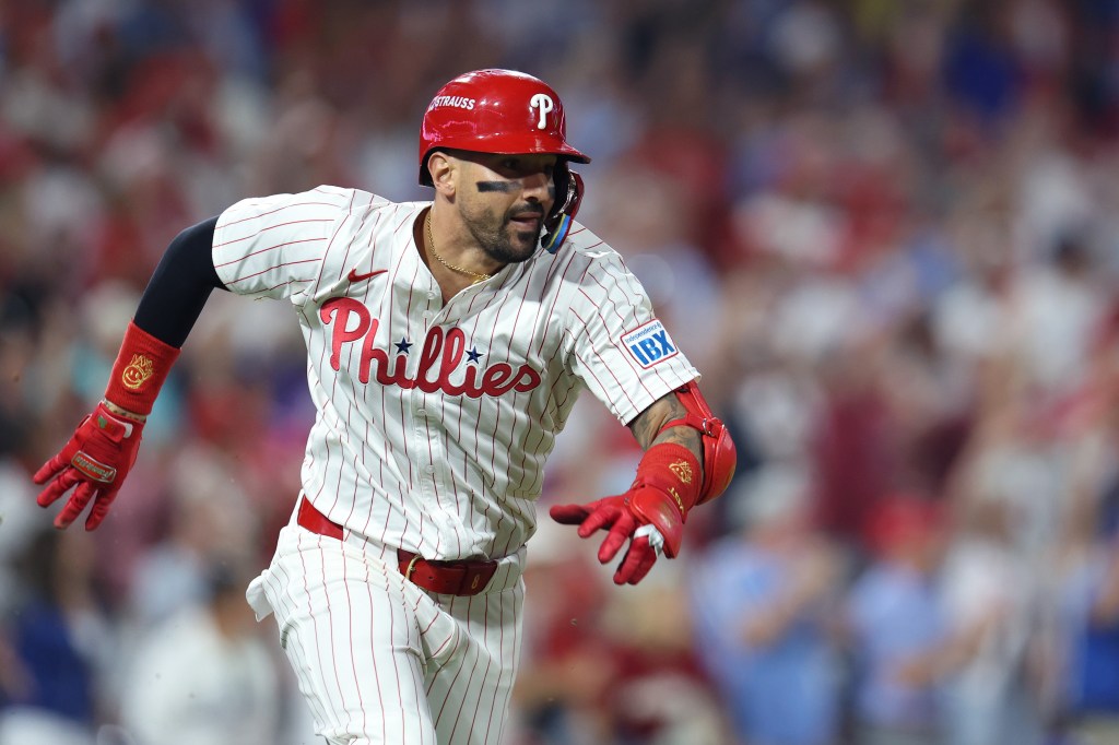 A baseball player in a white Phillies jersey, red helmet, and red gloves is running on the field.