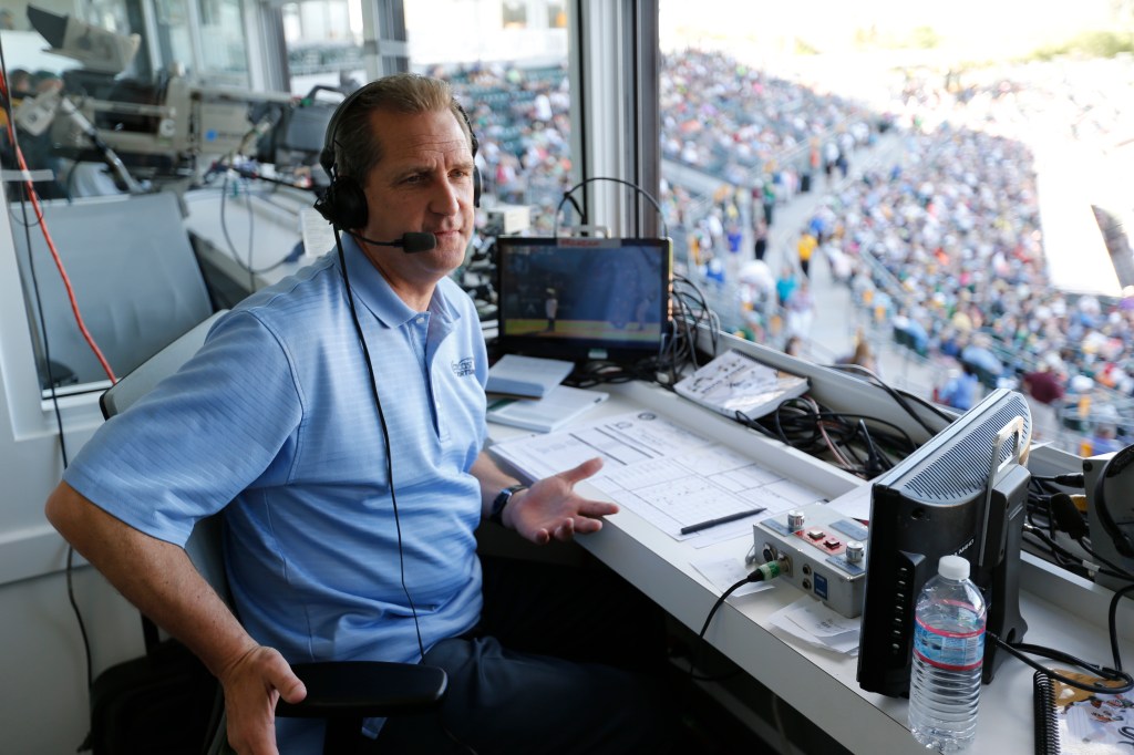 A man in a light blue polo shirt and headset, sitting in a broadcast booth, gestures while looking to his right.