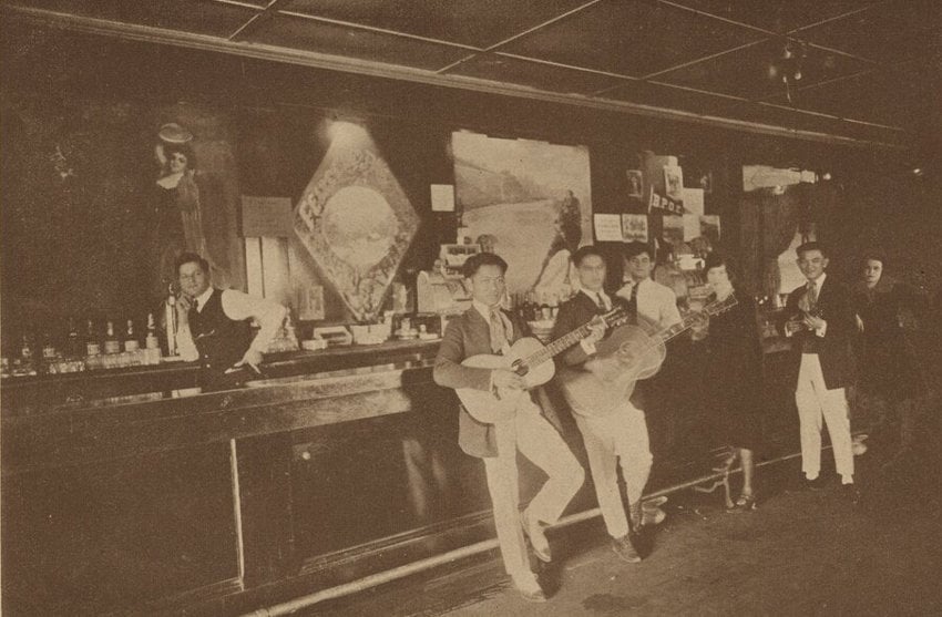 Sepia toned photo from 1922 of a candid moment at the Vernon Bar in Tijuana. Mexican guitar players are standing by a bar with American clientele in the background.