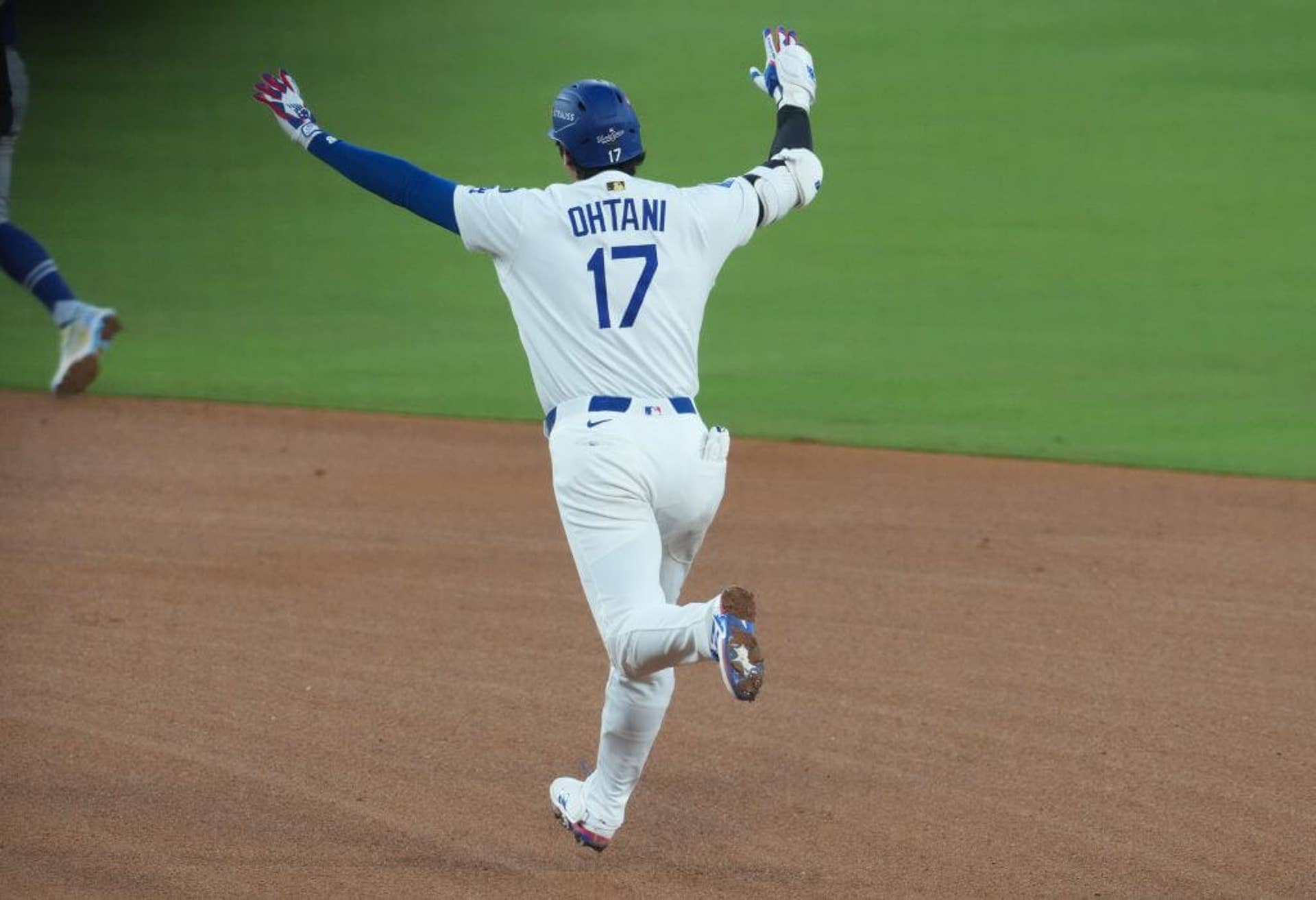 Shohei Ohtani, donning a white Dodgers uniform with a blue batting helmet and shown from behind, raises both arms in the air as he jogs around the bases after hitting a home run.