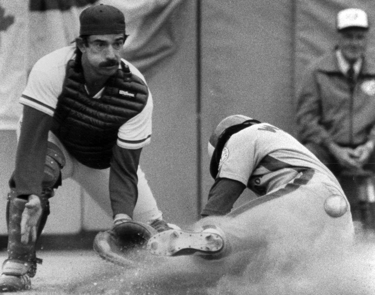 A black-and-white photo shows a back catcher blocking home plate as another player slides head first into the plate as the ball flies behind him.