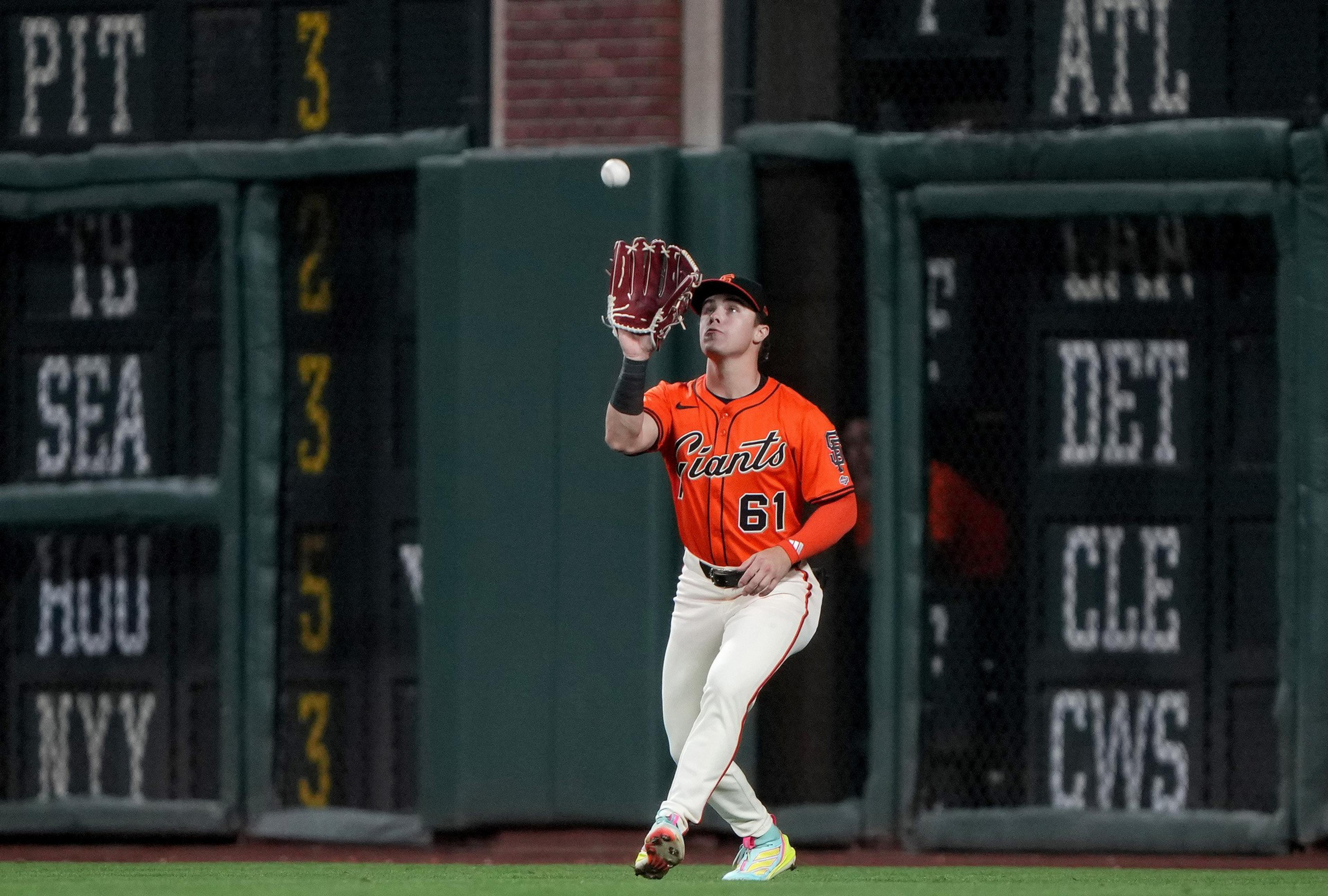 A baseball player in an orange Giants jersey with number 61 is preparing to catch a ball while standing on the field near the outfield wall.