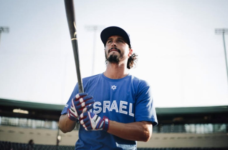 A man holding a baseball bat stands in the forefront of the frame, with a field and stands seen in the background. The man's jersey features the word "Israel."