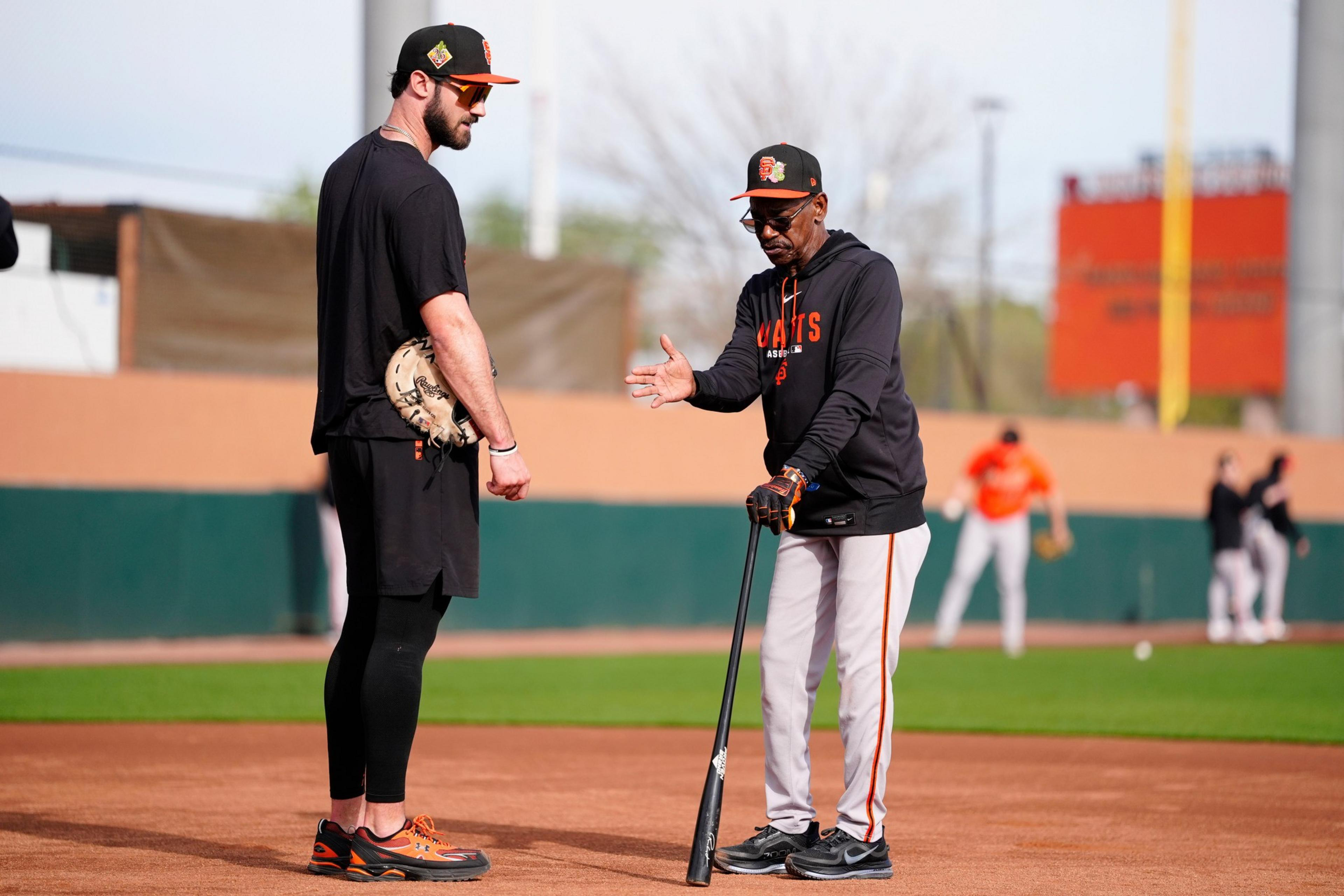 A baseball coach in black and gray is instructing a tall player dressed in black, holding a mitt, on a baseball field during practice.