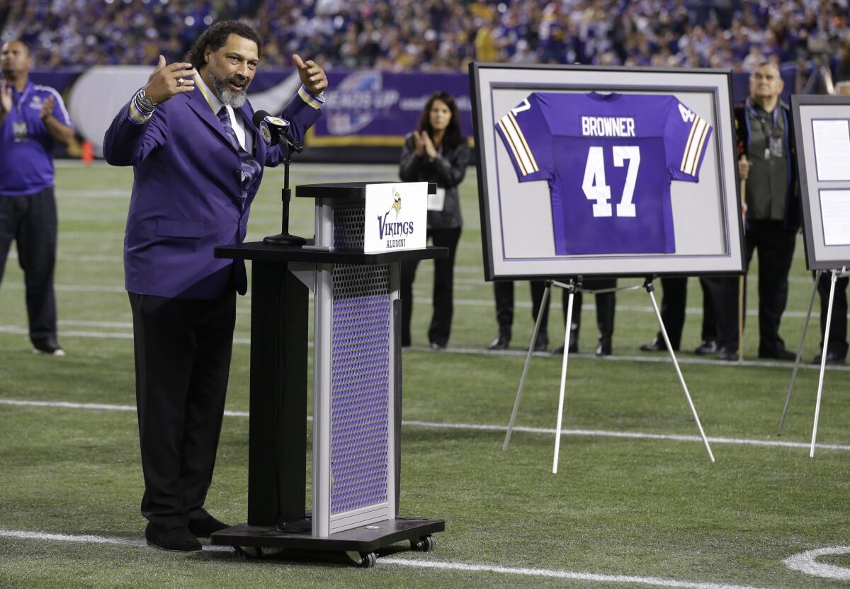 Joey Browner wears a purple blazer and speaks at a podium on a football field next to a framed Vikings jersey with his name